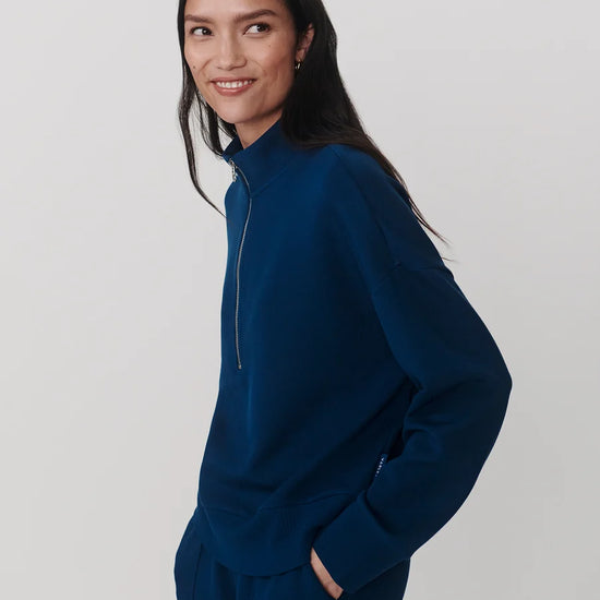 A woman wearing a dark blue mock-neck zip-up sweatshirt and matching sweatpants, photographed against a light gray studio background.