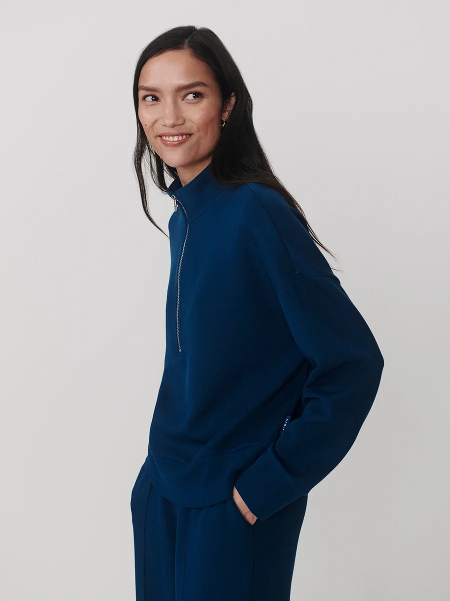 A woman wearing a dark blue mock-neck zip-up sweatshirt and matching sweatpants, photographed against a light gray studio background.