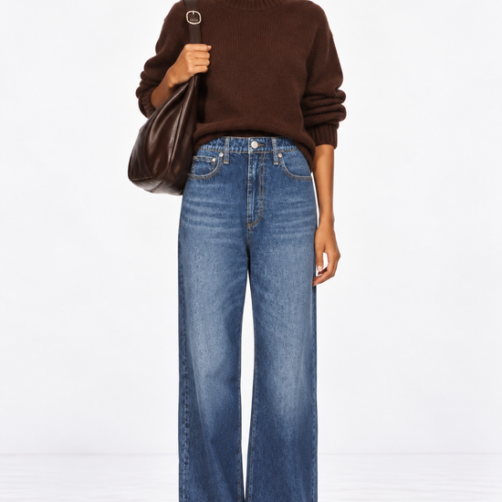 A model stands against a white studio background wearing a dark brown crewneck sweater, wide-leg blue jeans, and brown shoes, while carrying a dark brown leather shoulder bag.