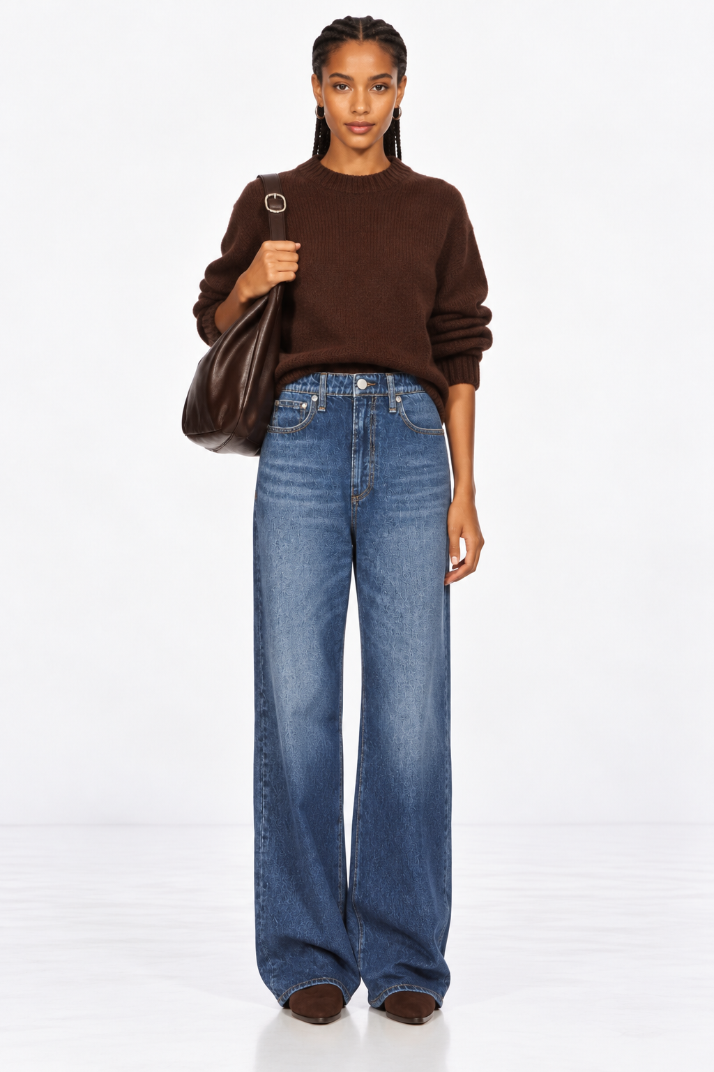 A model stands against a white studio background wearing a dark brown crewneck sweater, wide-leg blue jeans, and brown shoes, while carrying a dark brown leather shoulder bag.