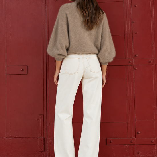 A woman stands with her back to the camera wearing a beige fuzzy bubble-sleeve sweater and white wide-leg pants, posing in front of a large red metal door.