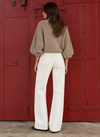 A woman stands with her back to the camera wearing a beige fuzzy bubble-sleeve sweater and white wide-leg pants, posing in front of a large red metal door.