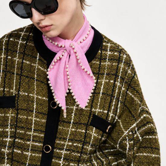 A close-up of a model wearing a pink corduroy neck scarf with pearl trim, styled with a green and black plaid tweed jacket and black sunglasses against a light gray background.