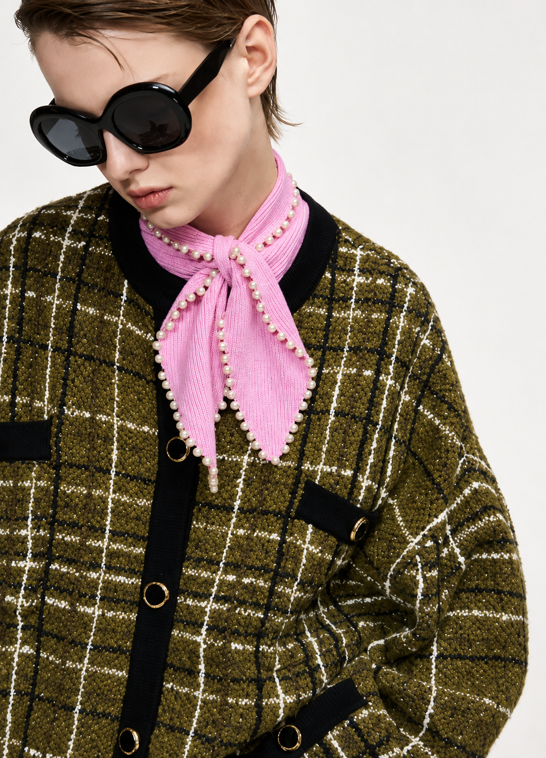 A close-up of a model wearing a pink corduroy neck scarf with pearl trim, styled with a green and black plaid tweed jacket and black sunglasses against a light gray background.