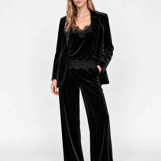 A woman modeling a black velvet pantsuit with a matching blazer and wide-leg trousers, paired with a black lace-trimmed top and black pointed heels, against a plain white studio background.