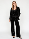 A woman modeling a black velvet pantsuit with a matching blazer and wide-leg trousers, paired with a black lace-trimmed top and black pointed heels, against a plain white studio background.