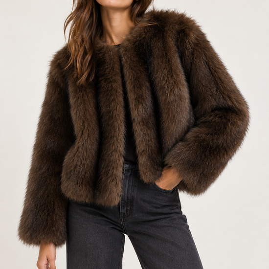 A woman modeling a cropped, dark brown faux fur jacket with a wide collar, paired with dark gray jeans, against a plain light gray studio background.