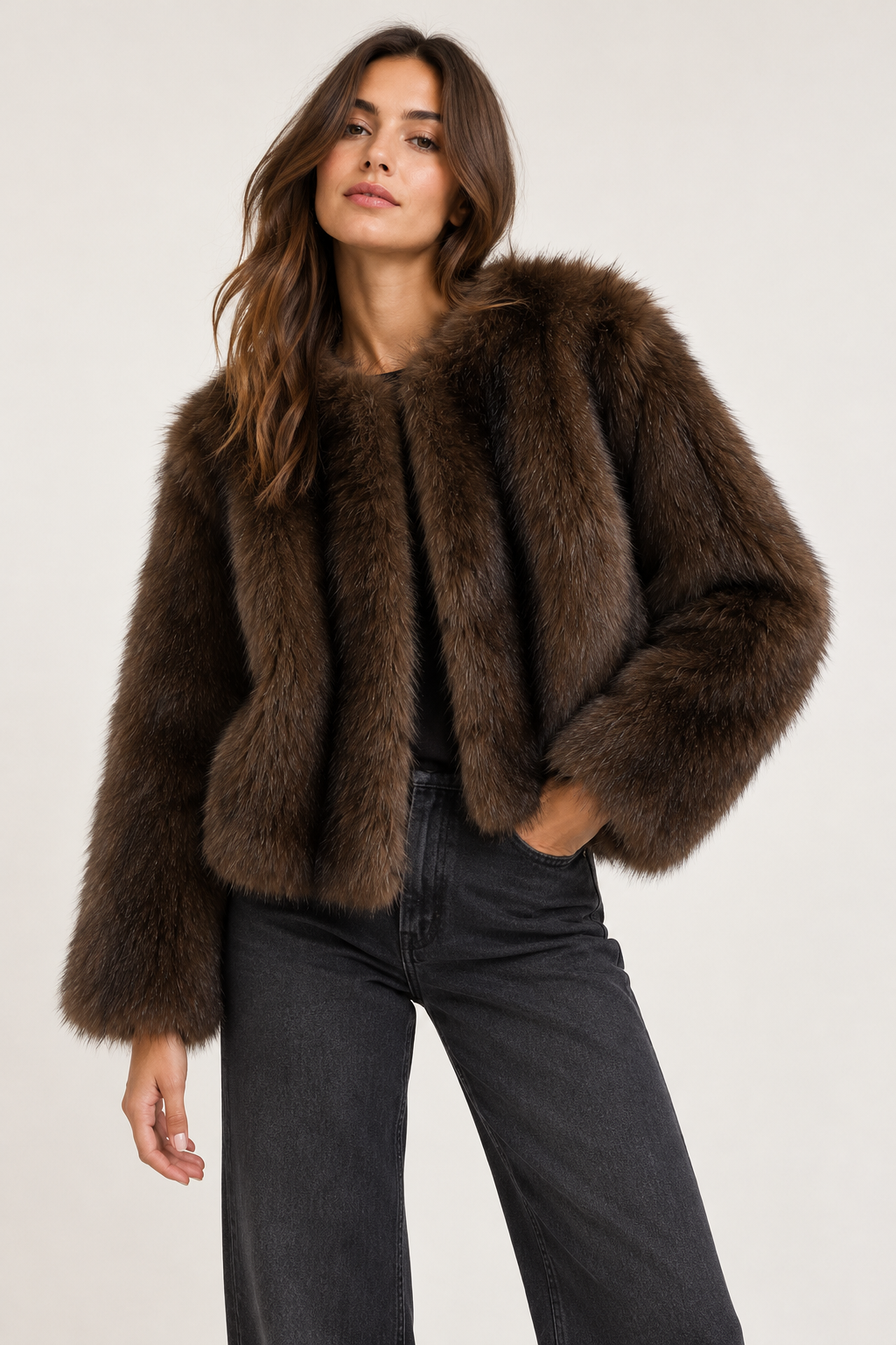 A woman modeling a cropped, dark brown faux fur jacket with a wide collar, paired with dark gray jeans, against a plain light gray studio background.