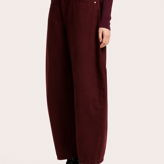 A model is shown from the waist down wearing high-waisted, wide-leg trousers in a deep burgundy, styled with a bright pink belt, a matching burgundy long-sleeve top, and black pointed-toe ankle boots, against a plain off-white studio background.