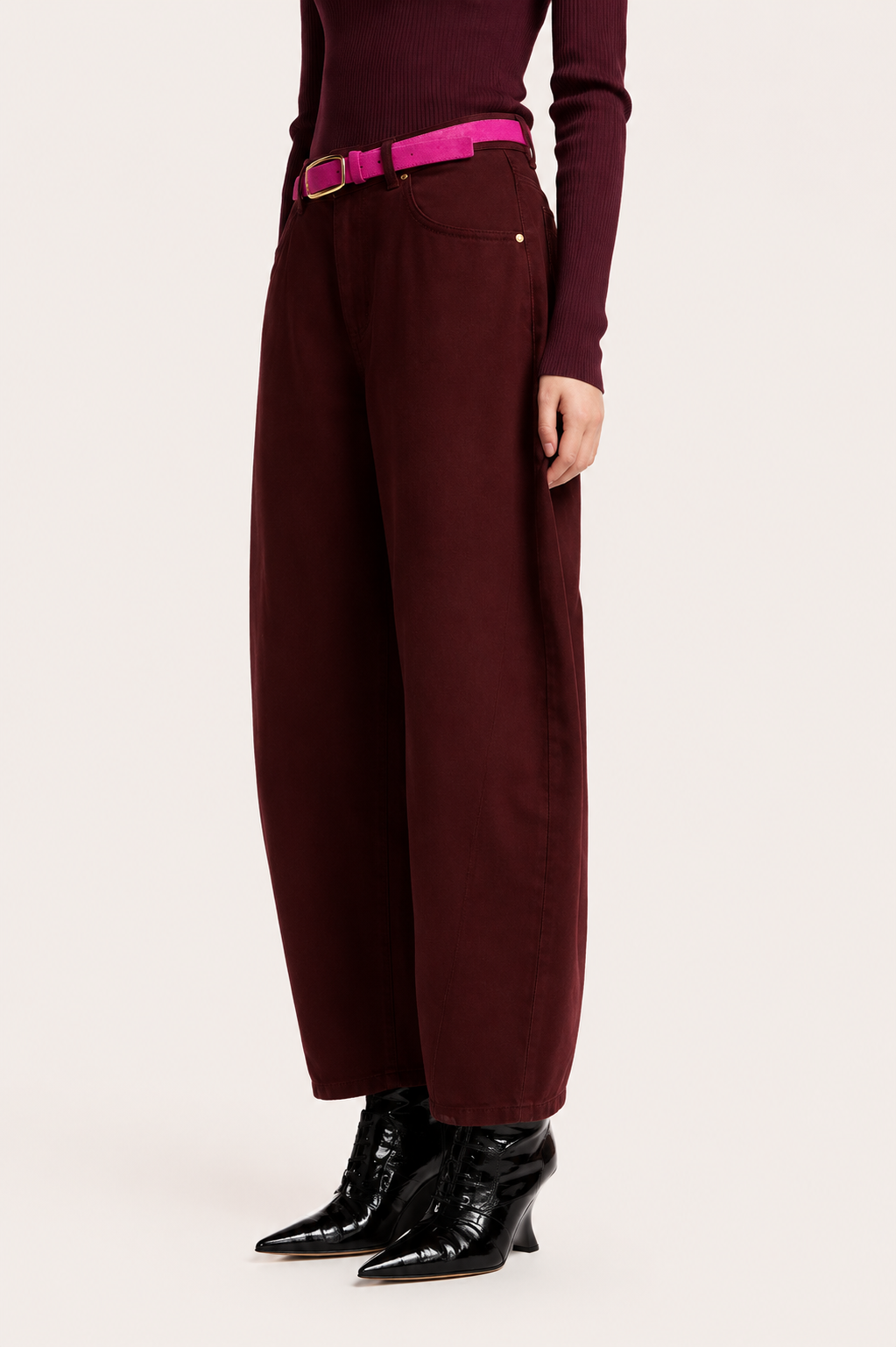 A model is shown from the waist down wearing high-waisted, wide-leg trousers in a deep burgundy, styled with a bright pink belt, a matching burgundy long-sleeve top, and black pointed-toe ankle boots, against a plain off-white studio background.