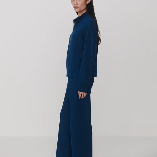 A model is shown in profile wearing a matching navy blue knit set consisting of a mock-neck long-sleeve top and wide-leg trousers, set against a plain light gray studio background.