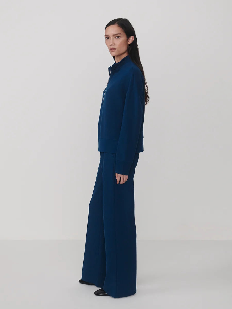 A model is shown in profile wearing a matching navy blue knit set consisting of a mock-neck long-sleeve top and wide-leg trousers, set against a plain light gray studio background.