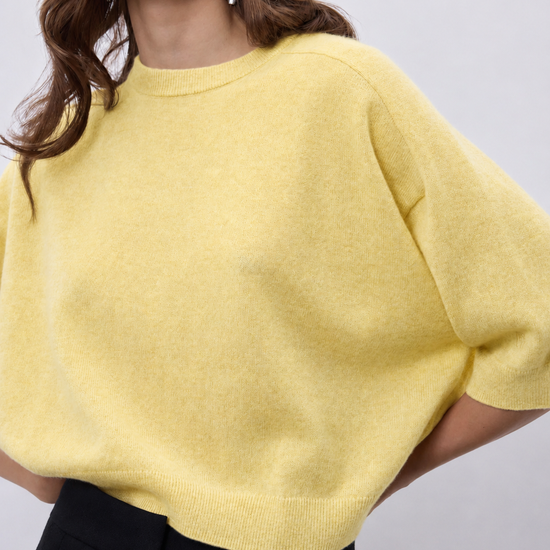 A close-up of a woman wearing a pale yellow knit sweater with short dolman sleeves and black trousers, set against a plain light gray background.