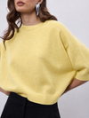 A close-up of a woman wearing a pale yellow knit sweater with short dolman sleeves and black trousers, set against a plain light gray background.