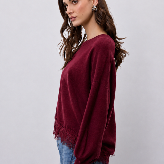 A woman wearing a long-sleeve crewneck sweater in deep burgundy with a lace hem detail, paired with blue jeans, against a light gray studio background.