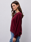 A woman wearing a long-sleeve crewneck sweater in deep burgundy with a lace hem detail, paired with blue jeans, against a light gray studio background.