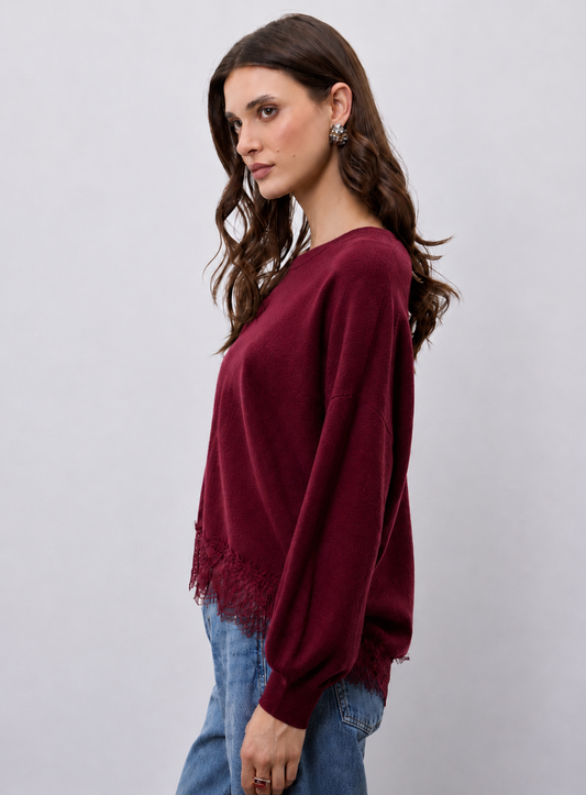 A woman wearing a long-sleeve crewneck sweater in deep burgundy with a lace hem detail, paired with blue jeans, against a light gray studio background.