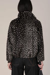 A person shown from the back wearing a dark cropped jacket with an allover light beige animal-like spot pattern, paired with dark jeans, against a plain light beige studio background.