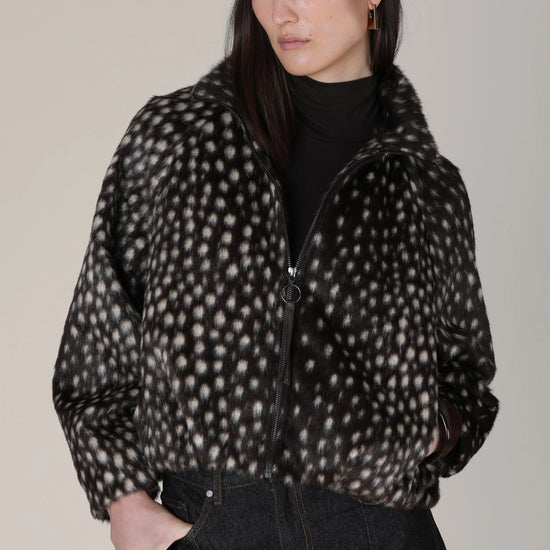A model wearing a black beret, a black turtleneck, and a black-and-white spotted zip-up jacket, paired with dark jeans, against a plain light gray studio background.