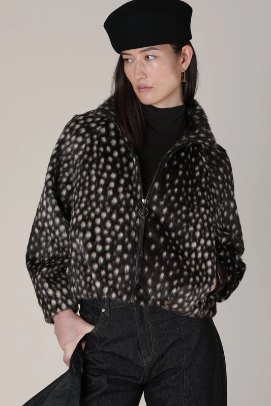 A model wearing a black beret, a black turtleneck, and a black-and-white spotted zip-up jacket, paired with dark jeans, against a plain light gray studio background.