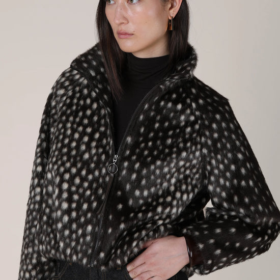 A model wearing a black beret, a black turtleneck, and a black fuzzy cropped jacket with a white spotted pattern, paired with dark jeans against a plain beige background.