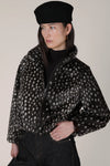 A model wearing a black beret, a black turtleneck, and a black fuzzy cropped jacket with a white spotted pattern, paired with dark jeans against a plain beige background.