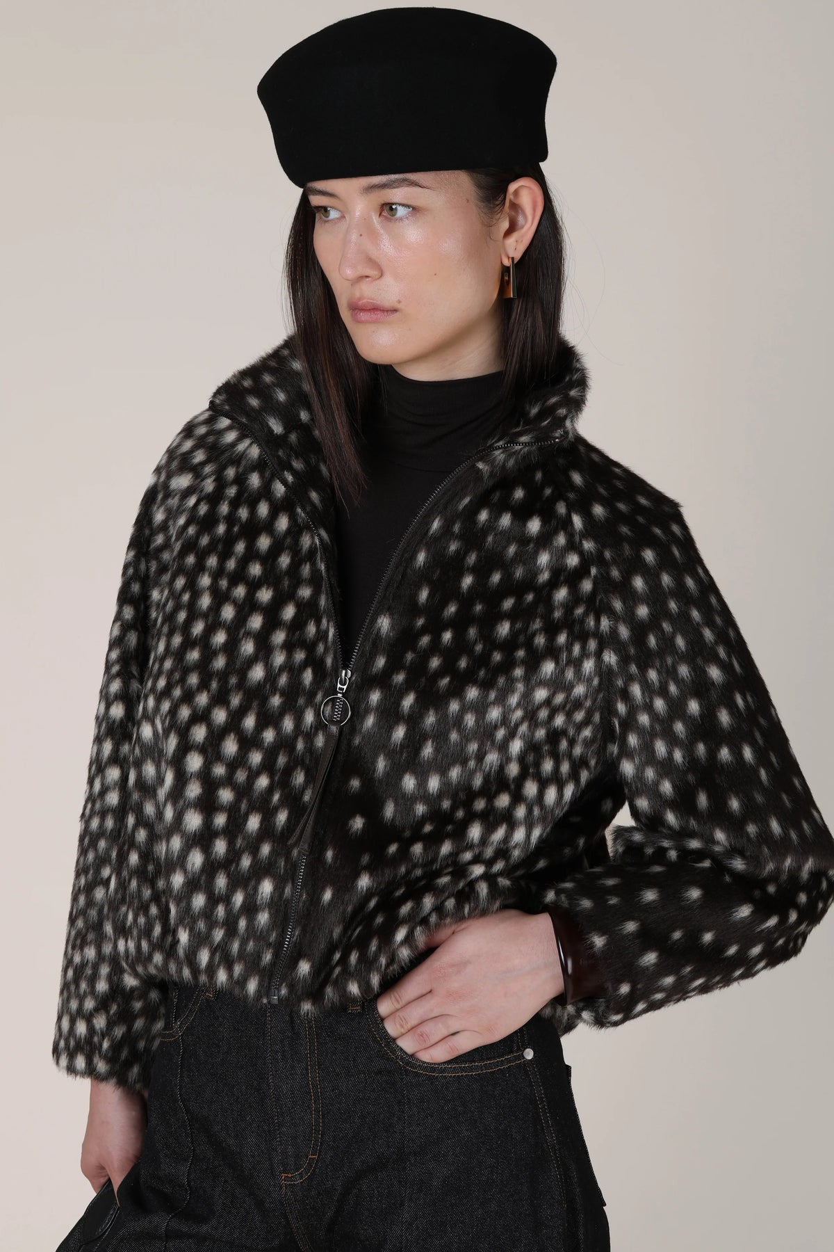 A model wearing a black beret, a black turtleneck, and a black fuzzy cropped jacket with a white spotted pattern, paired with dark jeans against a plain beige background.