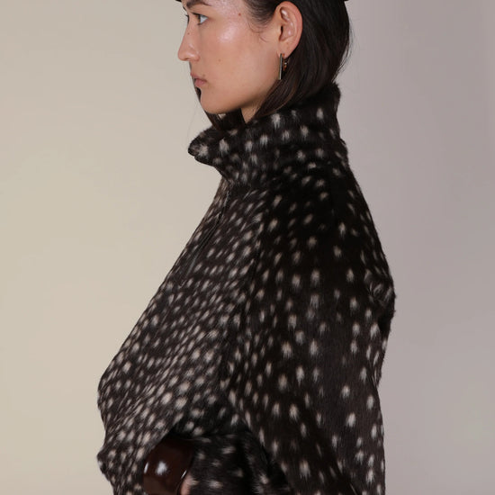 A side-profile view of a model wearing a black felt beret and a black faux-fur jacket with a white spotted pattern, set against a plain light beige background.
