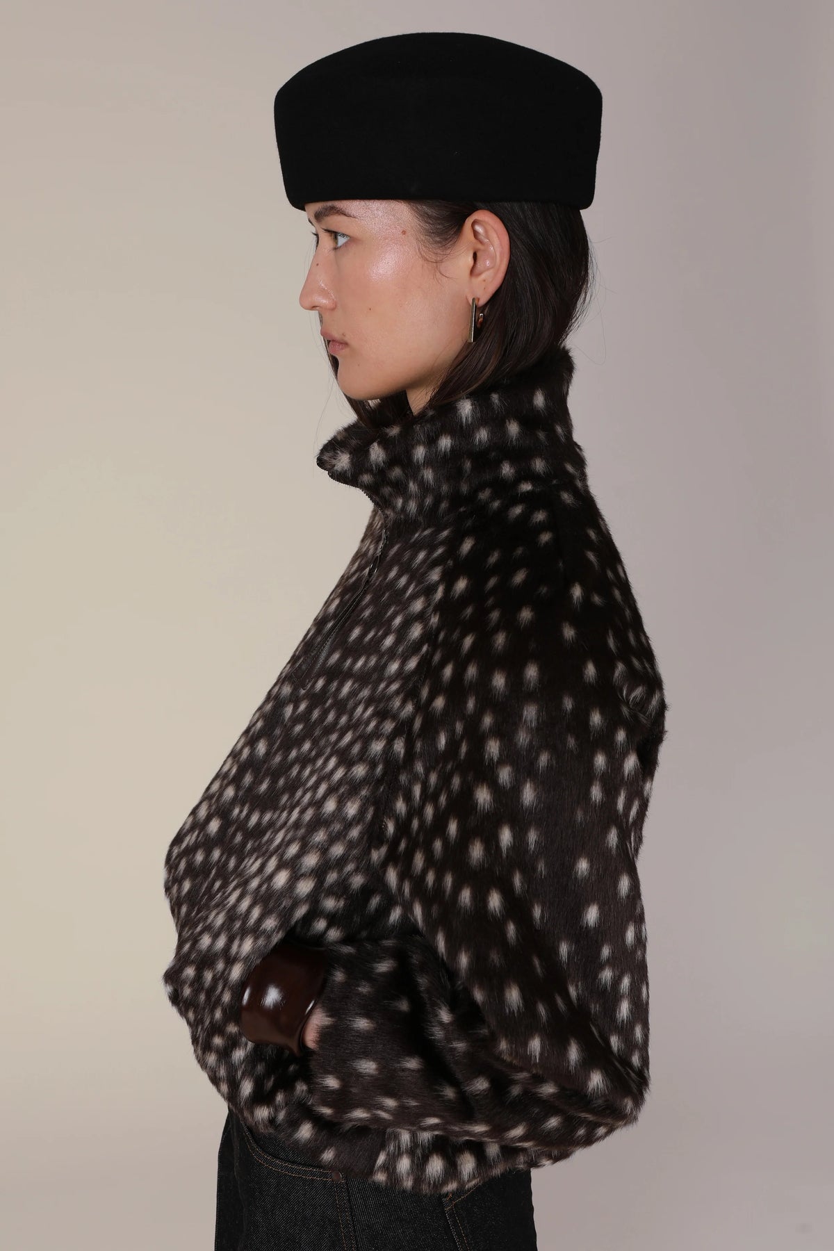 A side-profile view of a model wearing a black felt beret and a black faux-fur jacket with a white spotted pattern, set against a plain light beige background.