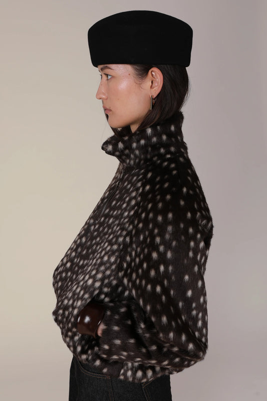 A side-profile view of a model wearing a black felt beret and a black faux-fur jacket with a white spotted pattern, set against a plain light beige background.
