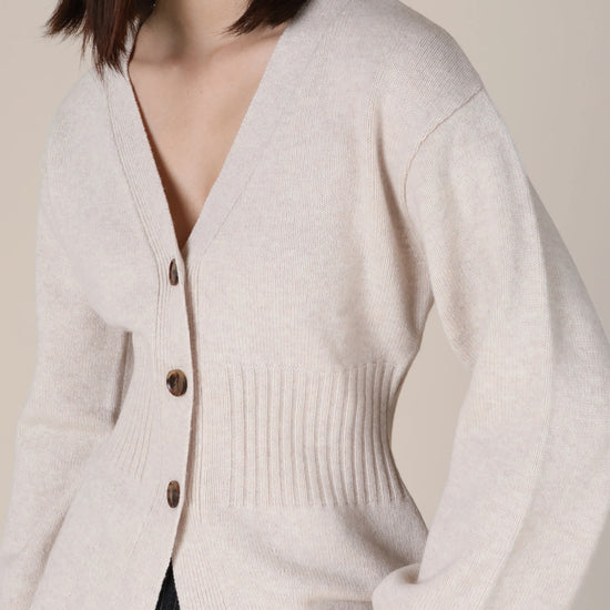 A woman wearing a light beige knit cardigan with dark brown buttons, shown from the chest down against a plain beige background.