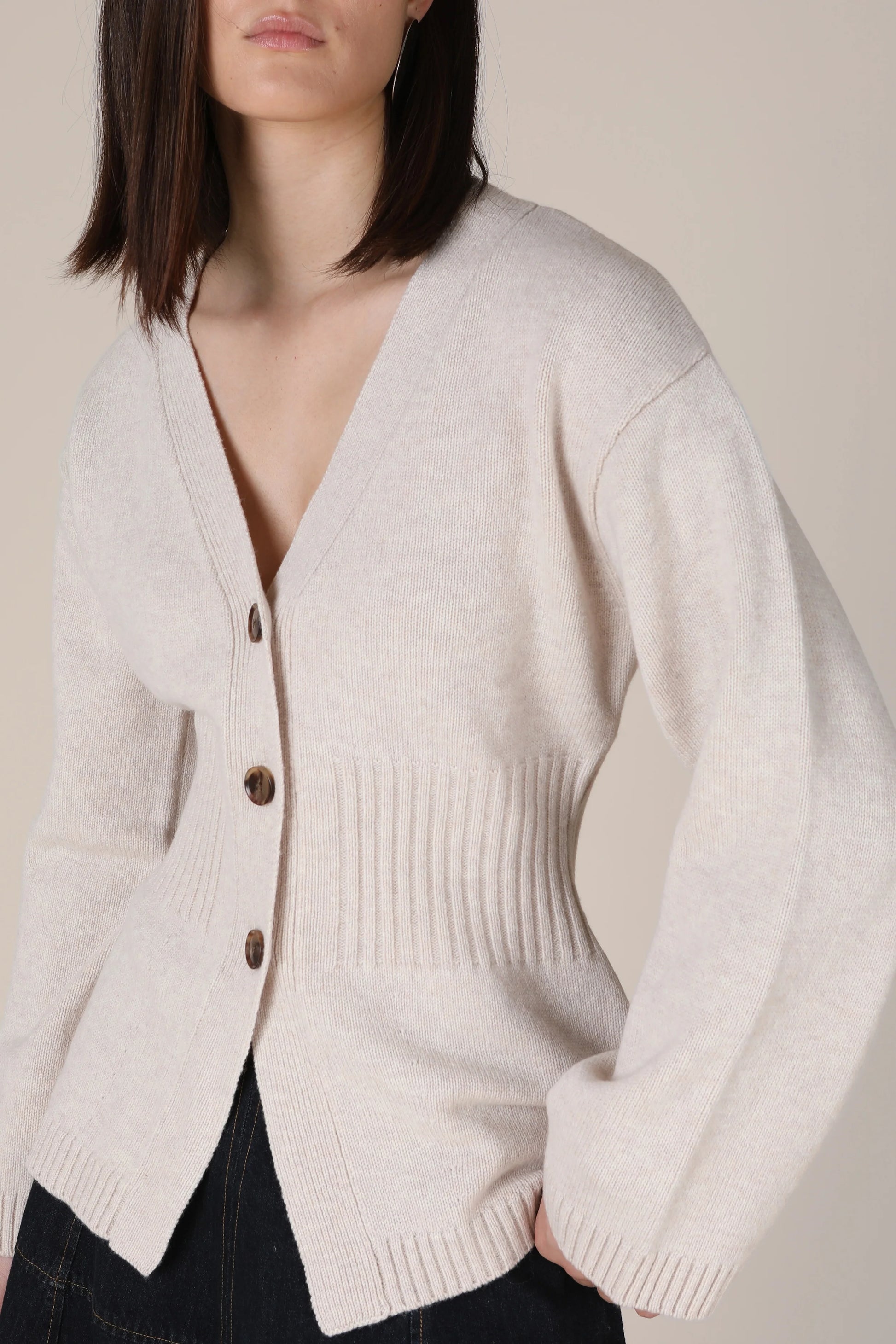 A woman wearing a light beige knit cardigan with dark brown buttons, shown from the chest down against a plain beige background.