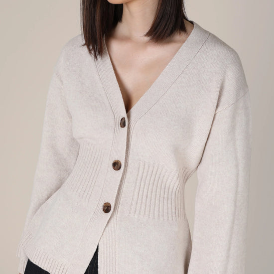 A woman wearing a light beige knit cardigan with brown buttons, paired with a dark skirt, against a solid beige background.