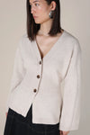 A woman wearing a light beige knit cardigan with brown buttons, paired with a dark skirt, against a solid beige background.