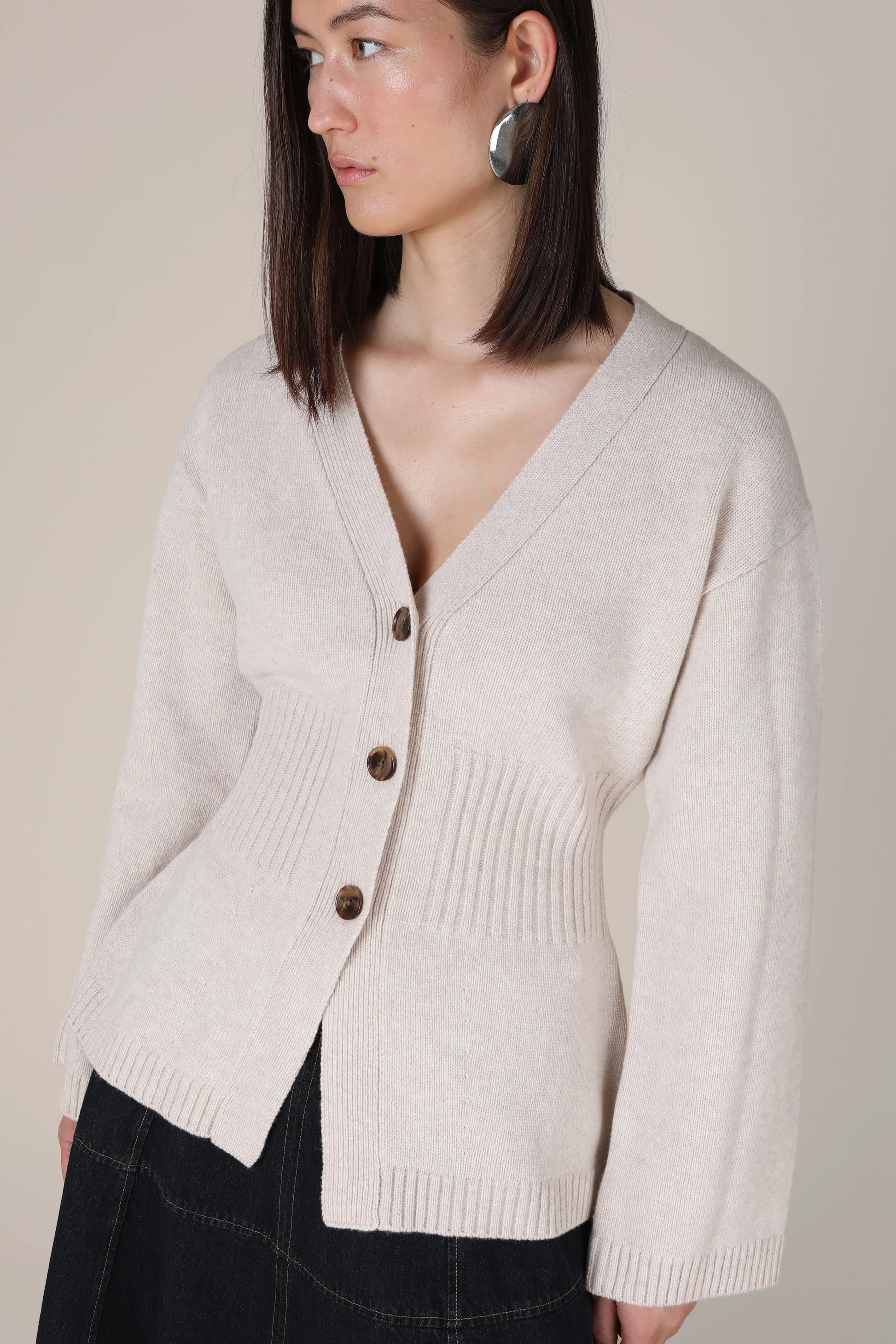 A woman wearing a light beige knit cardigan with brown buttons, paired with a dark skirt, against a solid beige background.