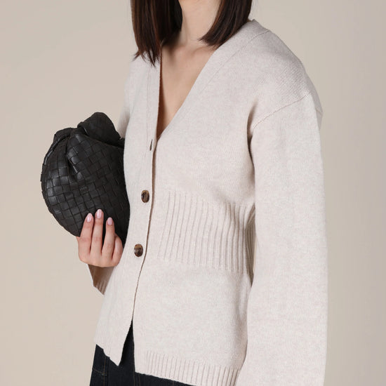 A woman wearing a light beige knit cardigan with dark buttons, paired with a dark skirt, holding a black woven clutch bag against a solid beige background.