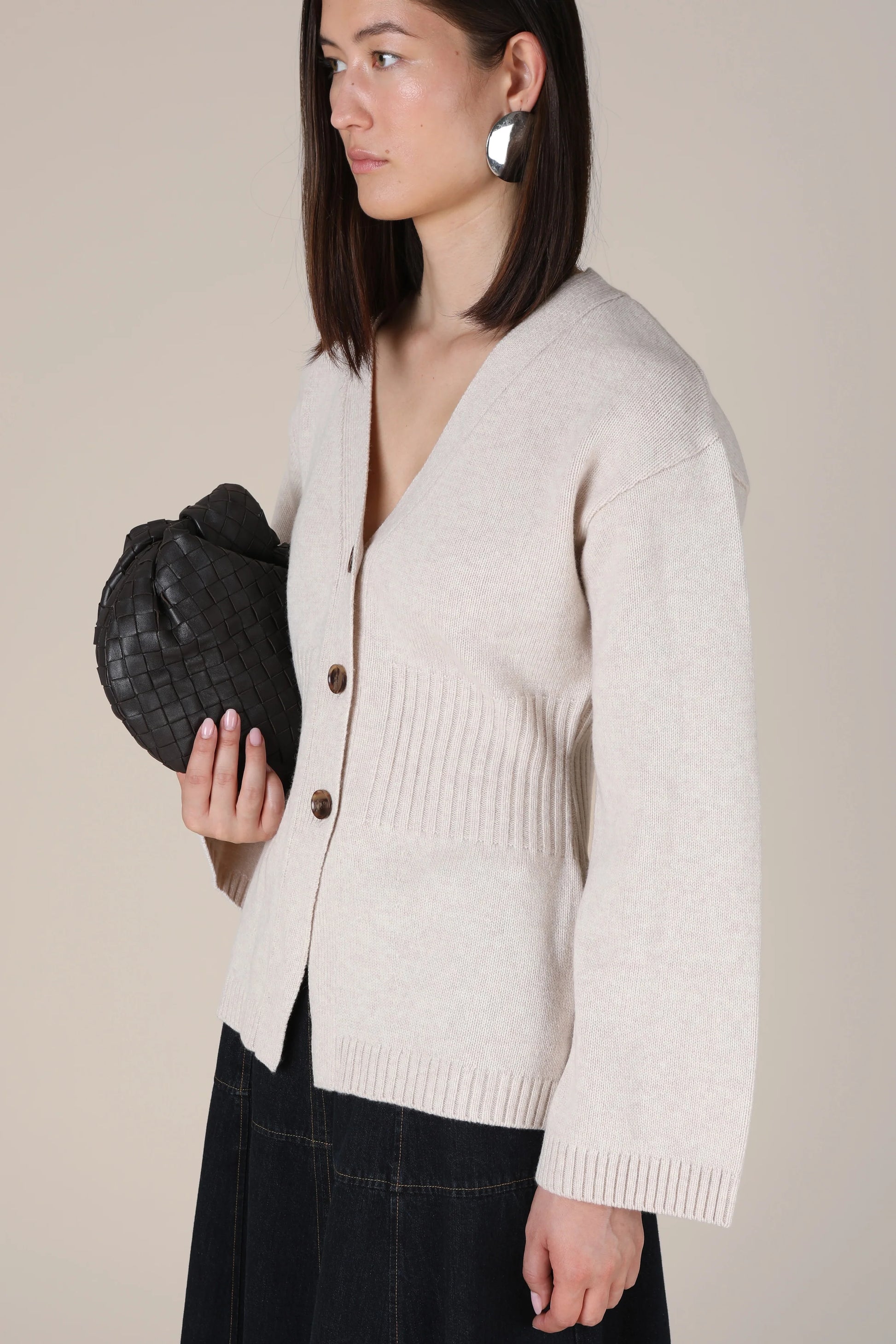 A woman wearing a light beige knit cardigan with dark buttons, paired with a dark skirt, holding a black woven clutch bag against a solid beige background.