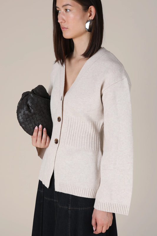 A woman wearing a light beige knit cardigan with dark buttons, paired with a dark skirt, holding a black woven clutch bag against a solid beige background.