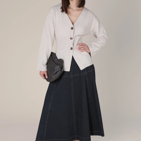A woman models a light beige ribbed cardigan with dark buttons, paired with a dark navy wide-leg skirt and a dark gray textured clutch bag, set against a plain beige studio background.