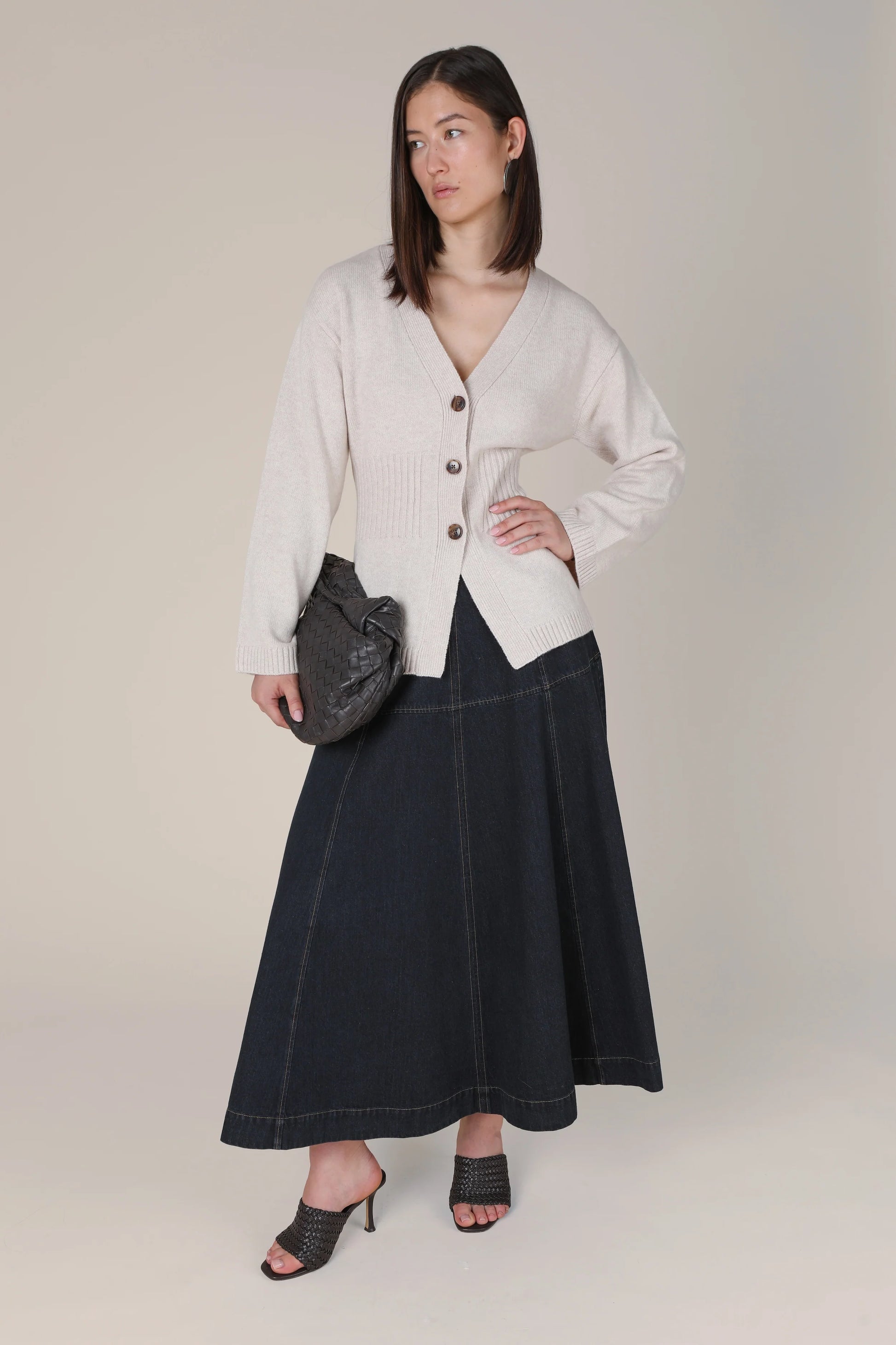 A woman models a light beige ribbed cardigan with dark buttons, paired with a dark navy wide-leg skirt and a dark gray textured clutch bag, set against a plain beige studio background.