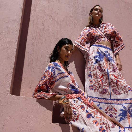Two women in colorful embroidered outfits standing against a beige wall.