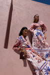 Two women in colorful embroidered outfits standing against a beige wall.
