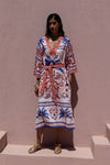 Woman wearing a colorful patterned dress standing against a pink wall.