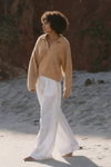 Person wearing a camel sweater and white pants standing on a sandy beach from Lilly Pilly