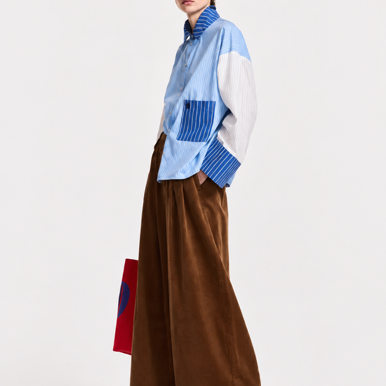 A model wearing a light blue and white color-blocked long-sleeve shirt with blue striped details, paired with wide-leg brown corduroy trousers, black sunglasses, and black shoes, holding a red and blue bag against a plain white studio background.
