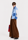 A model wearing a light blue and white color-blocked long-sleeve shirt with blue striped details, paired with wide-leg brown corduroy trousers, black sunglasses, and black shoes, holding a red and blue bag against a plain white studio background.