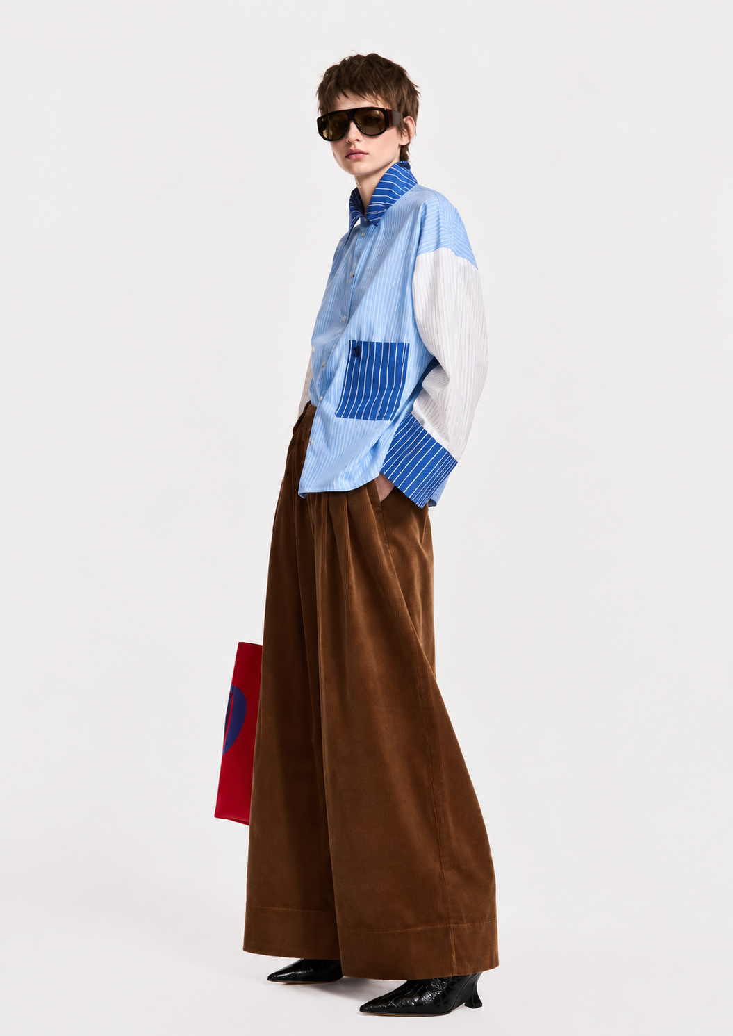 A model wearing a light blue and white color-blocked long-sleeve shirt with blue striped details, paired with wide-leg brown corduroy trousers, black sunglasses, and black shoes, holding a red and blue bag against a plain white studio background.