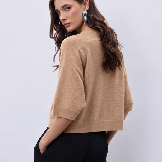 A woman wearing a cropped beige knit sweater with elbow-length sleeves and black wide-leg trousers, photographed against a light gray studio background.