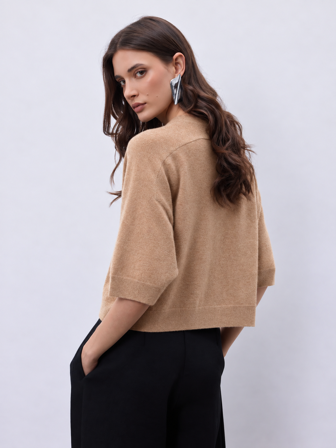 A woman wearing a cropped beige knit sweater with elbow-length sleeves and black wide-leg trousers, photographed against a light gray studio background.