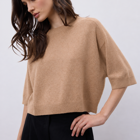 A woman wearing a cropped, short-sleeve knit sweater in a warm camel beige color, paired with black trousers, against a plain light gray studio background.